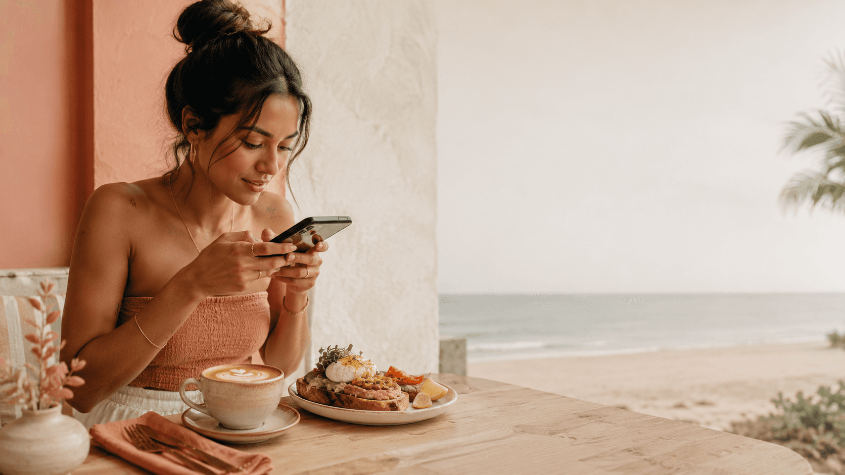 Café guest photographing her avocado toast and latte with her phone, with the beach in the background
